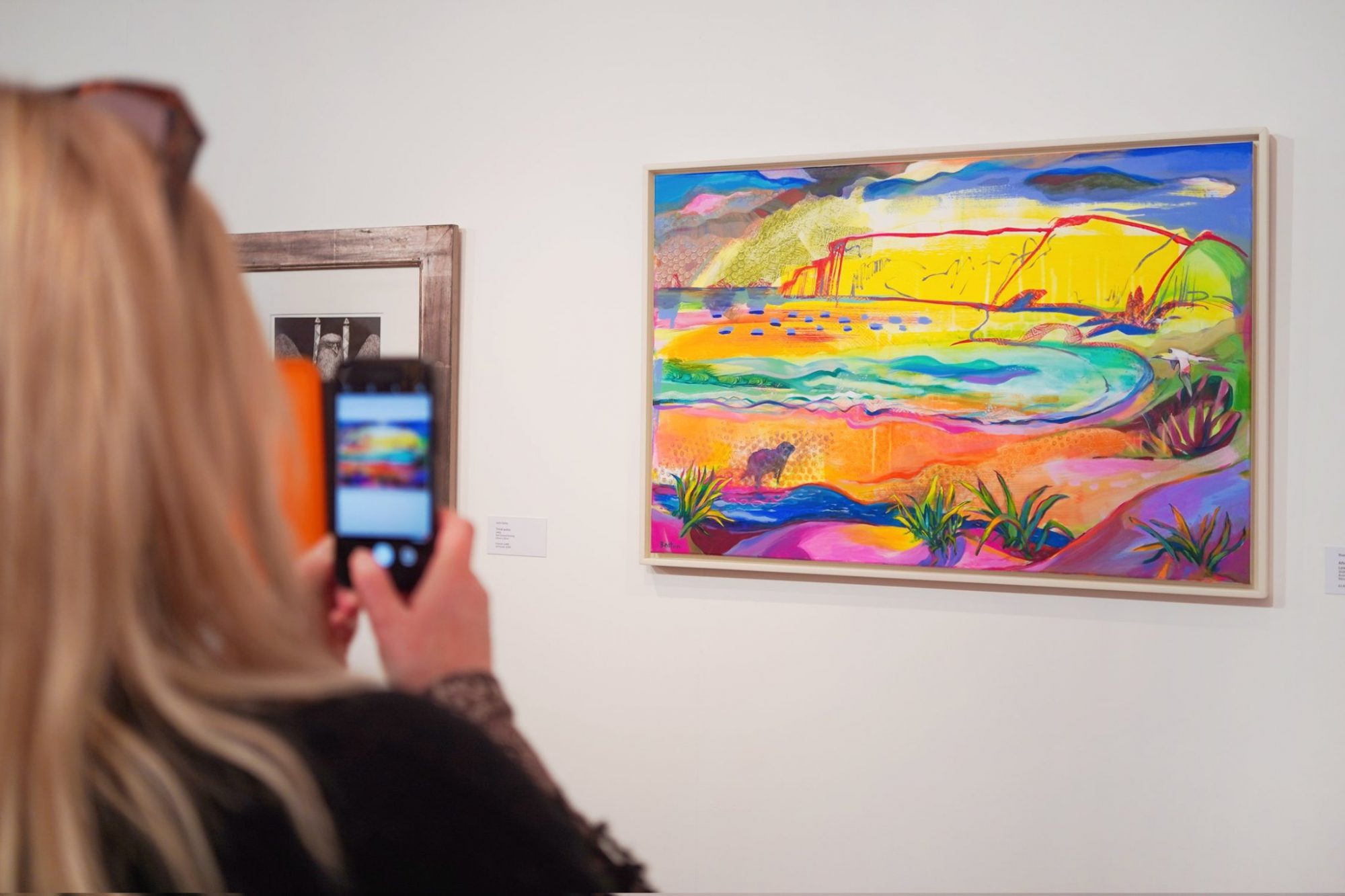 A woman stands in front of two paintings. She is holding her phone out in front of her taking a photograph of a colourful work of art of a landscape.